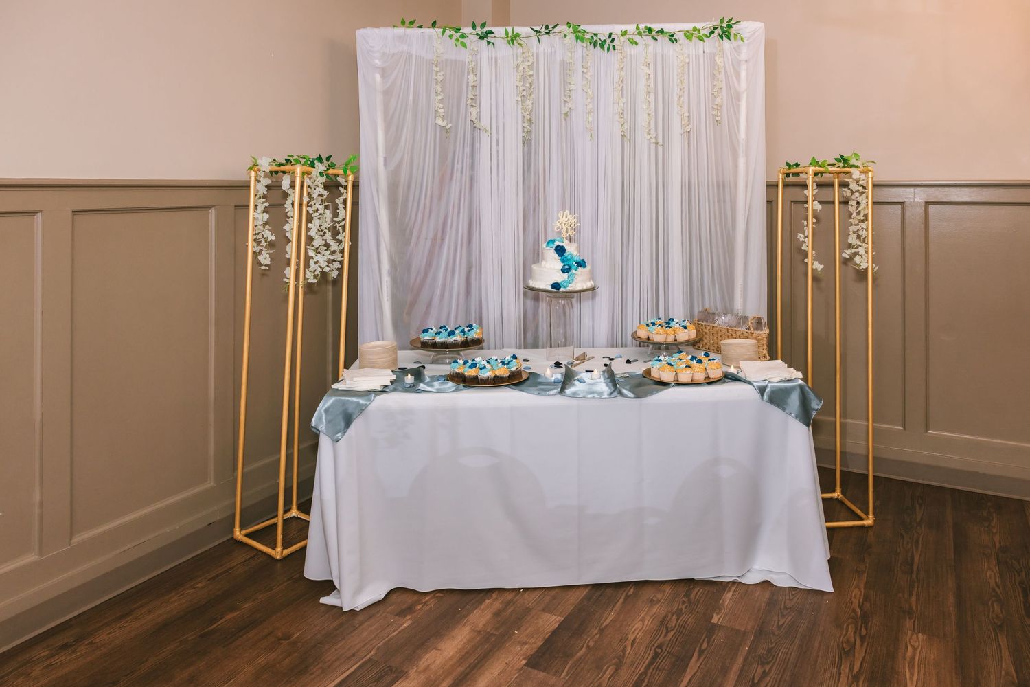 Elegant wedding dessert table with white draping, gold stands, blue floral accents, and a tiered cake centerpiece.