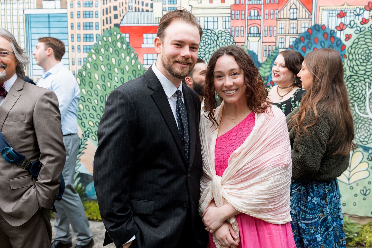 A man in a black suit and woman in pink dress with cream shawl smile at a colorful mural event.