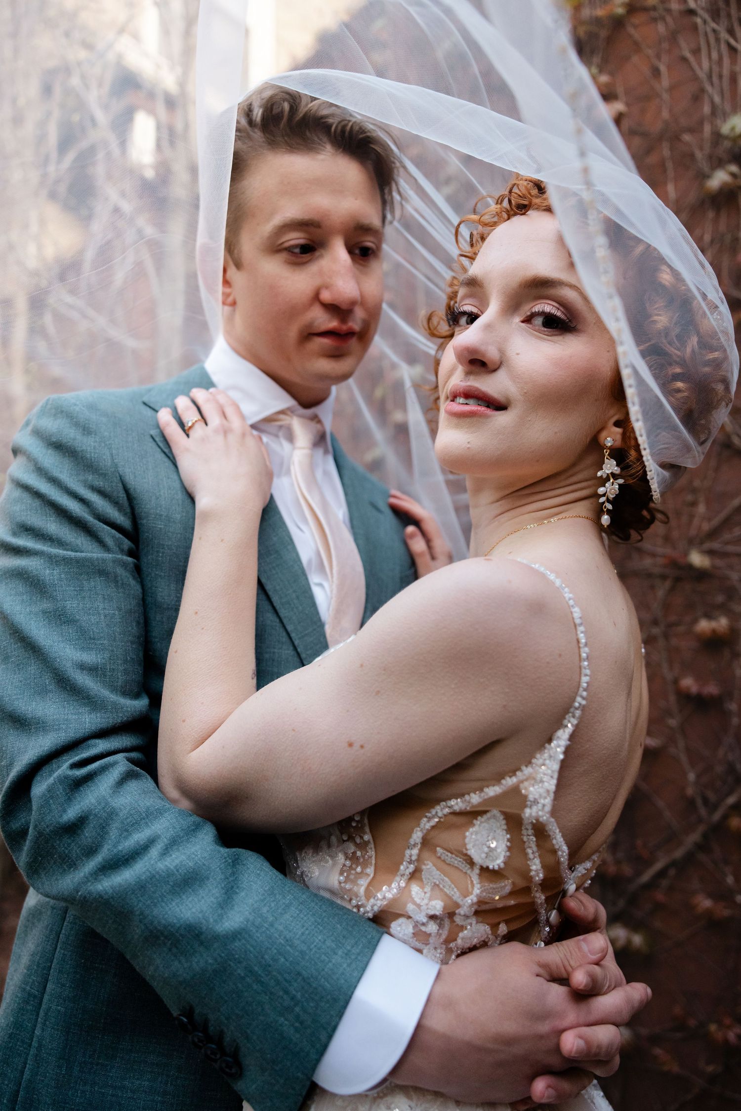 Bride and groom posing under wedding veil, bride in beaded gown with groom in teal suit embracing.