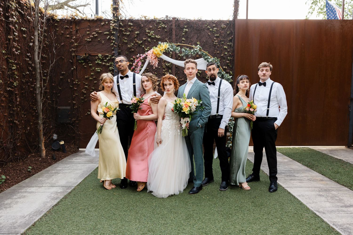 Wedding party stands together outdoors on green turf with a colorful floral arch backdrop on a bright day.