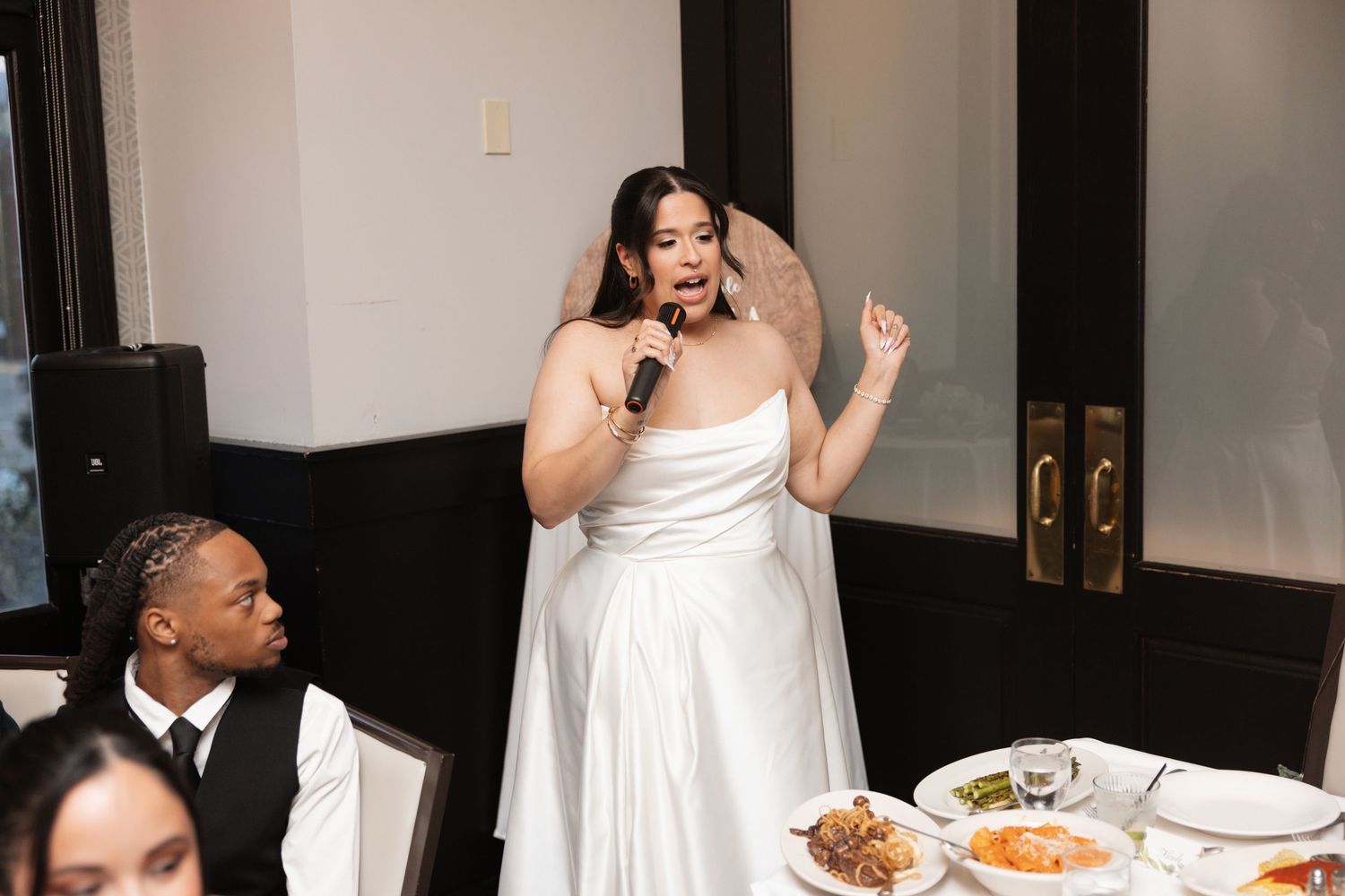 Bride in white gown speaking into microphone during wedding reception speech.