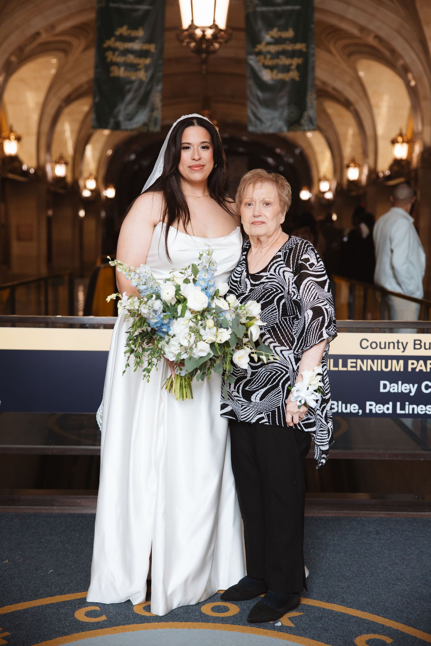 Bride in white gown holds blue and white bouquet posing with older woman in black patterned top at Chicago venue.