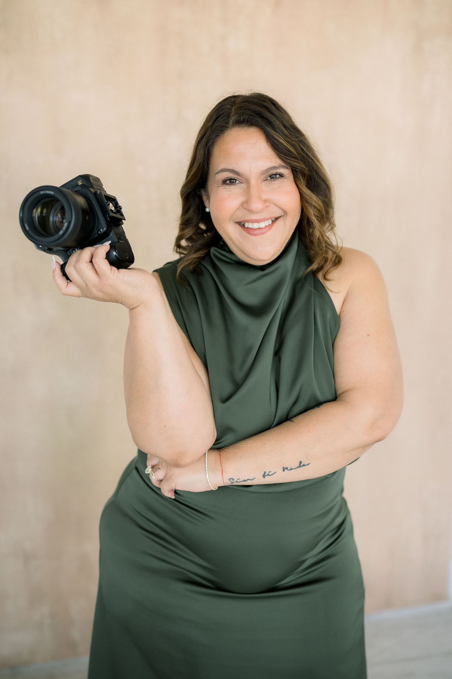 Professional photographer in green dress holding camera and smiling against neutral backdrop.