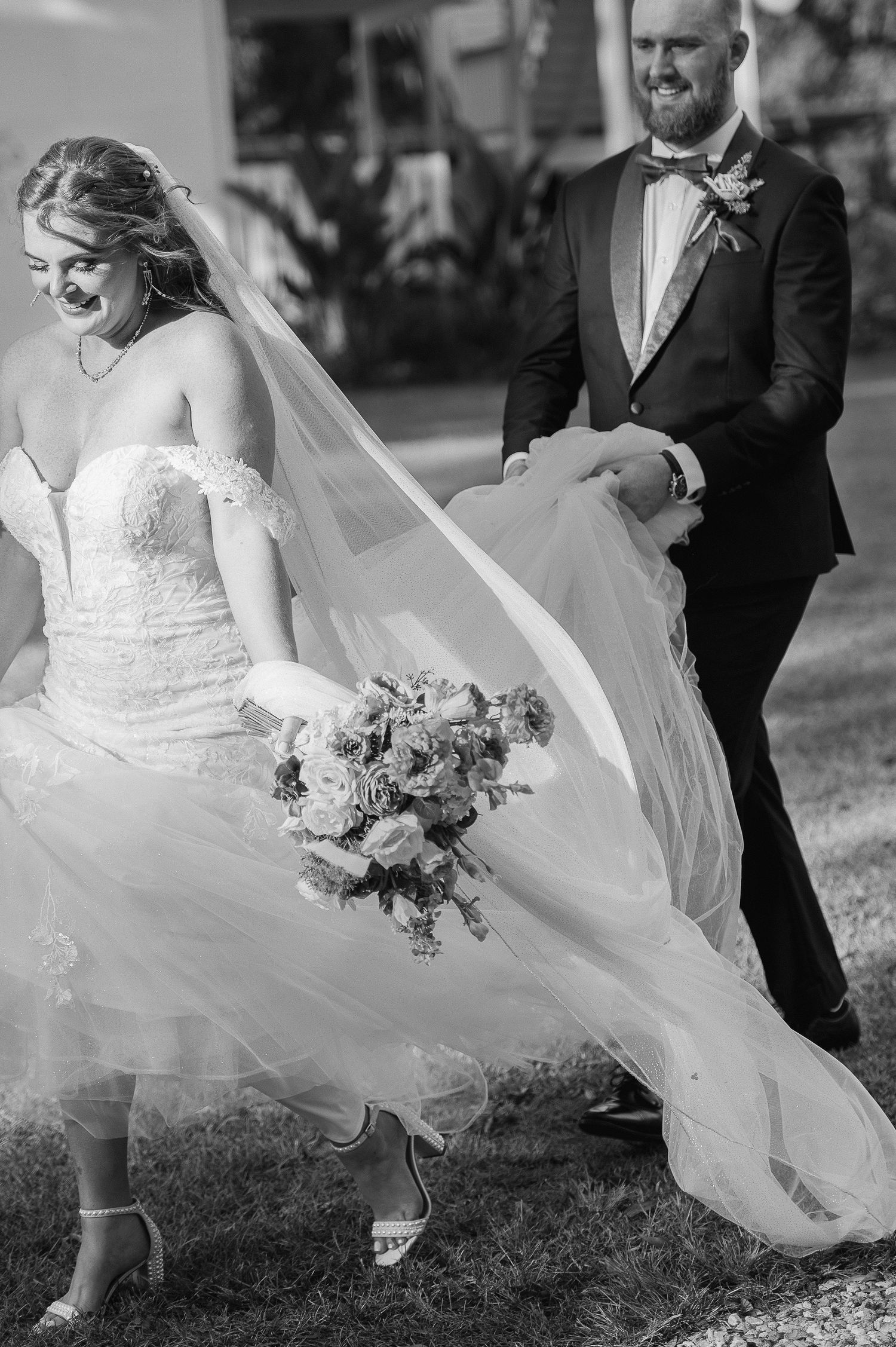 A bride in a white dress twirls outdoors as her veil flows in the wind while a groom looks on smiling.