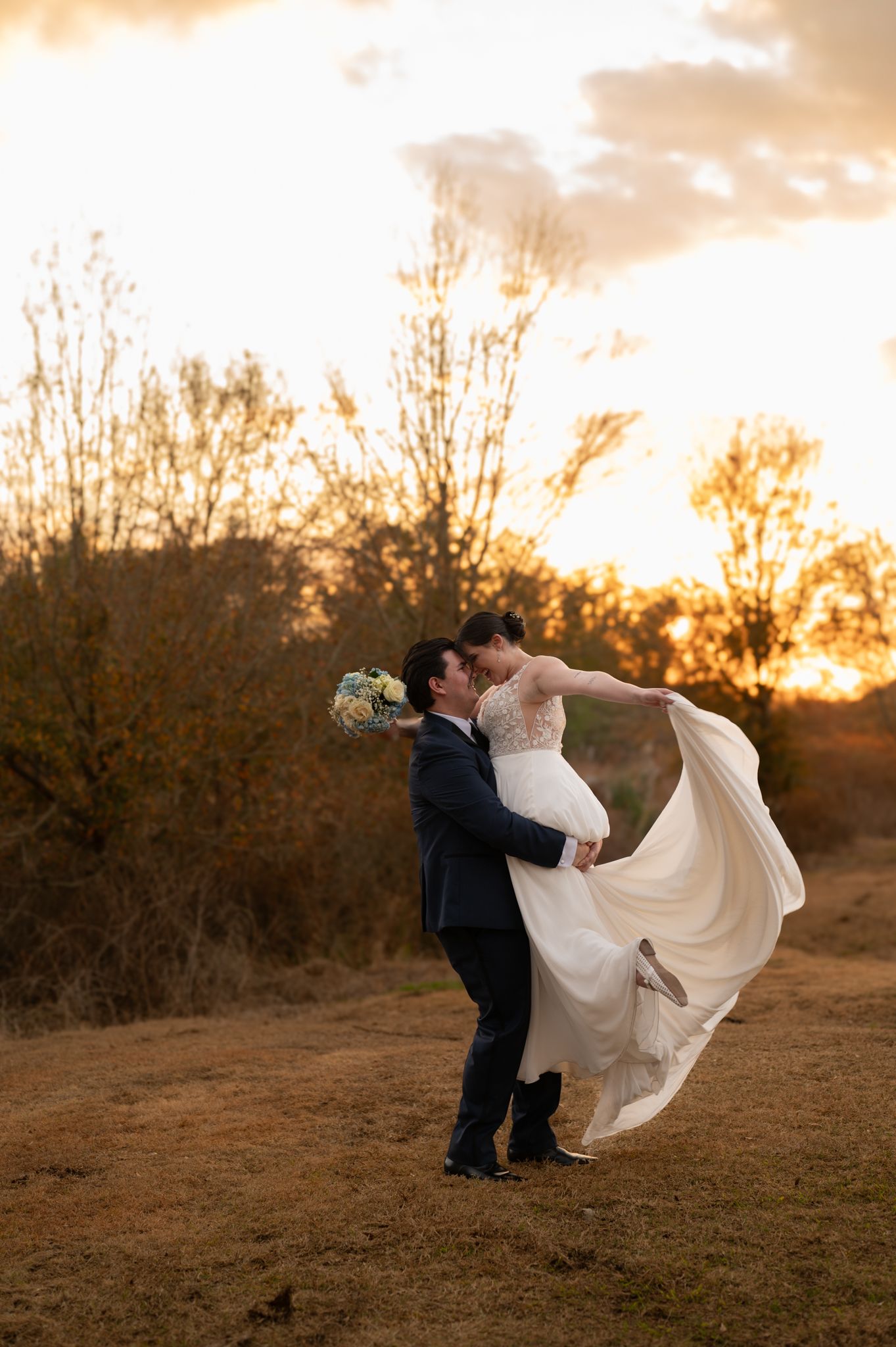 A romantic sunset silhouette of a couple in wedding attire embracing on a hilltop with flowing white dress and dramatic sky.