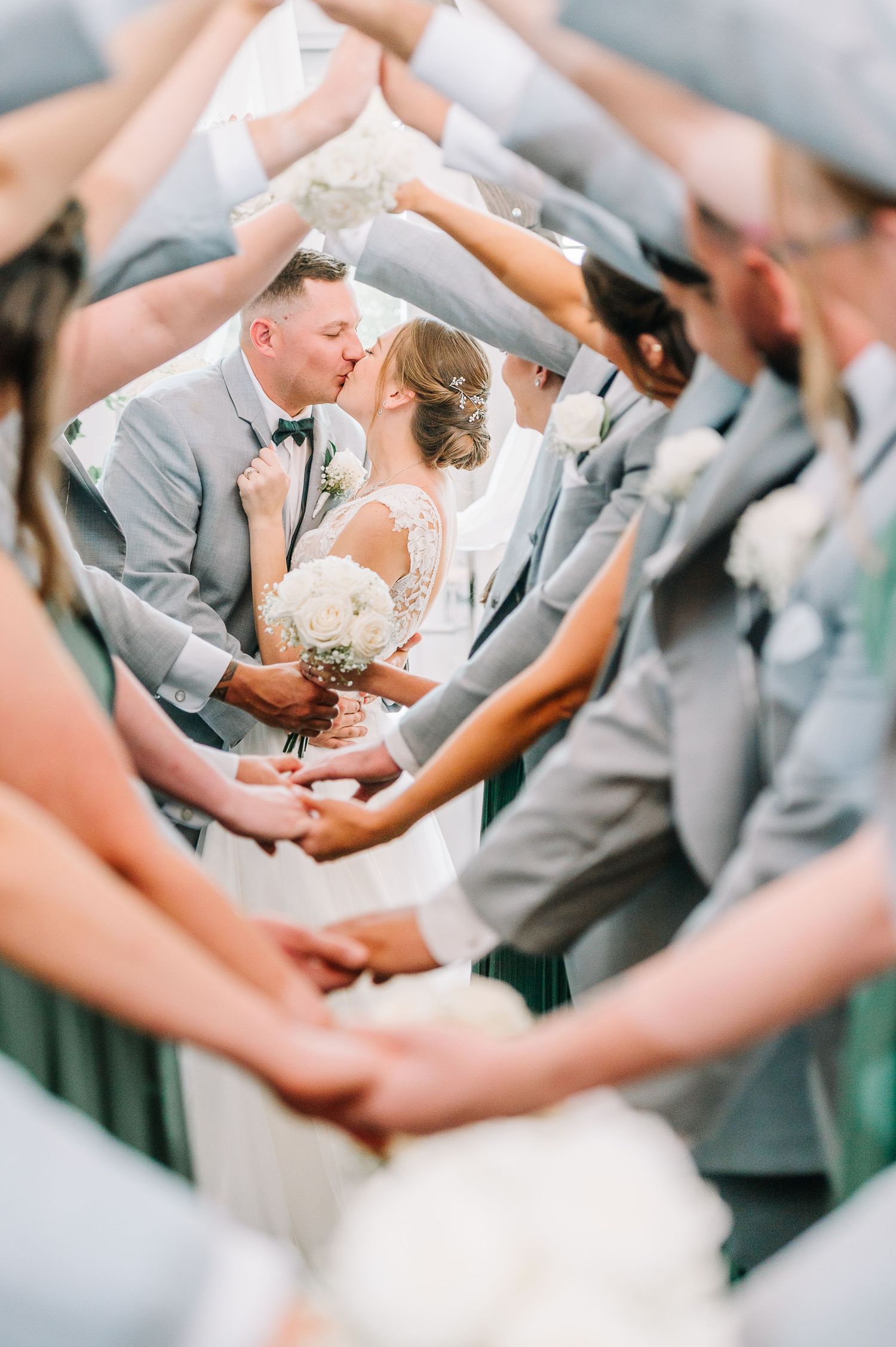Wedding guests hold up their hands in a circle formation while the newlyweds share a romantic kiss in the center.
