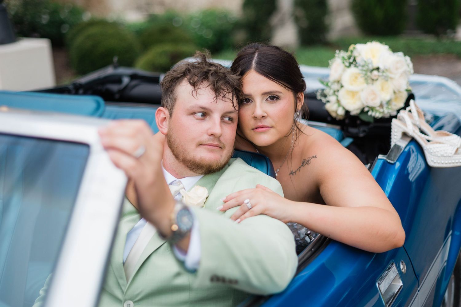 Bride and groom pose in a vintage blue convertible car, bride holding white bouquet, groom in sage green suit.