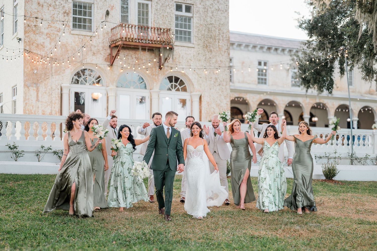 Wedding party walks together outside elegant venue, bride in white gown with groomsmen and bridesmaids in sage green dresses.