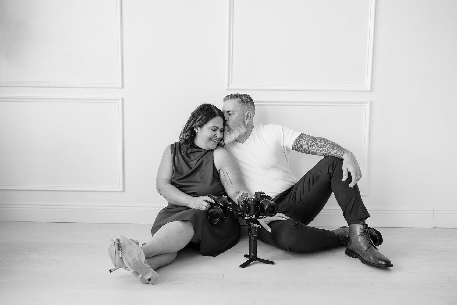 Black and white photo of a couple sitting on floor, leaning together tenderly, holding cameras in a bright studio.