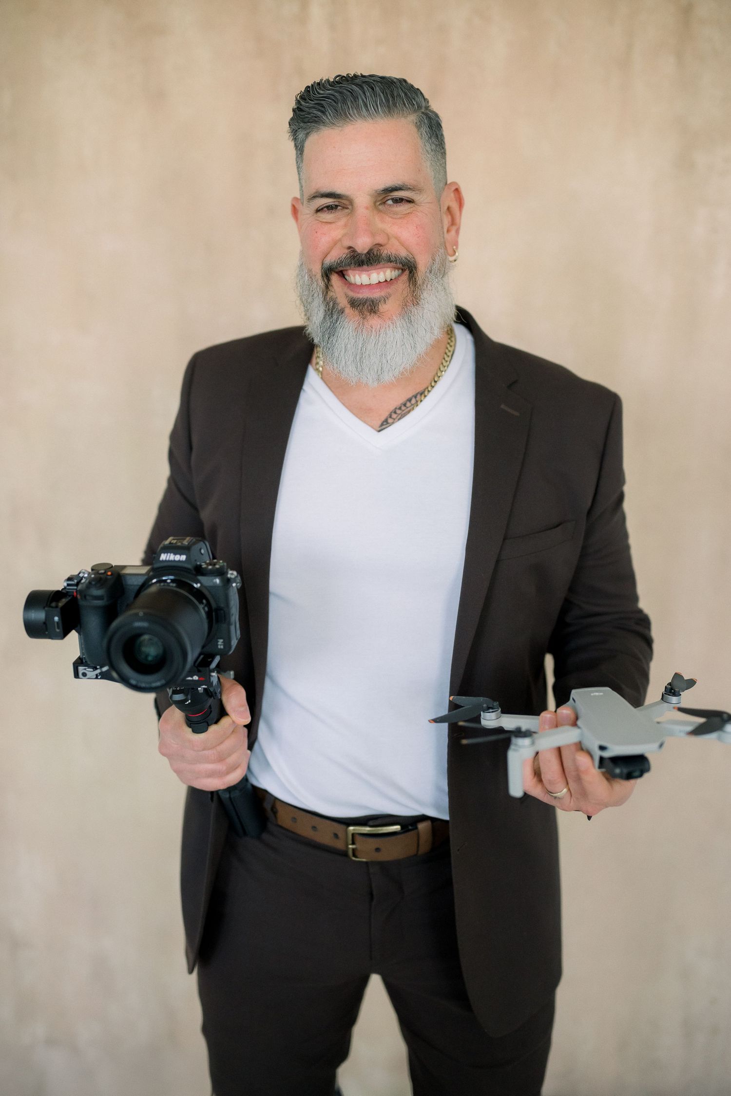 Smiling man with gray beard holding a Nikon camera on gimbal and drone, wearing black blazer and white V-neck shirt.
