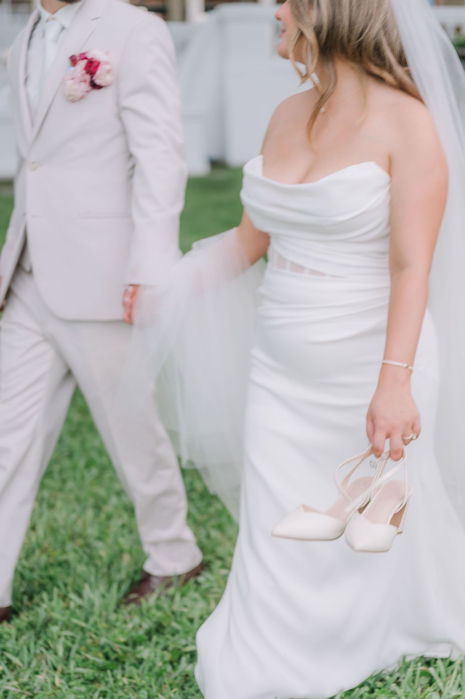 A bride in a white strapless gown holds her heels while standing next to groom in light beige suit with pink boutonniere.