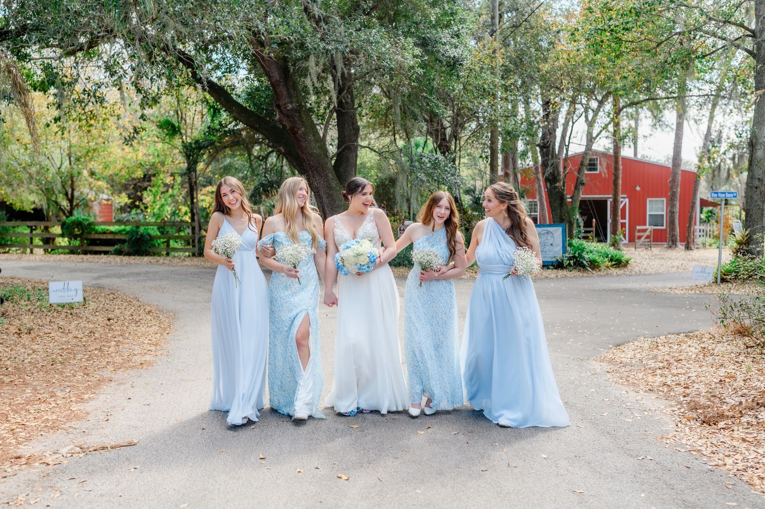 A group in light blue and white dresses walks together through a tree-lined pathway during an outdoor event.