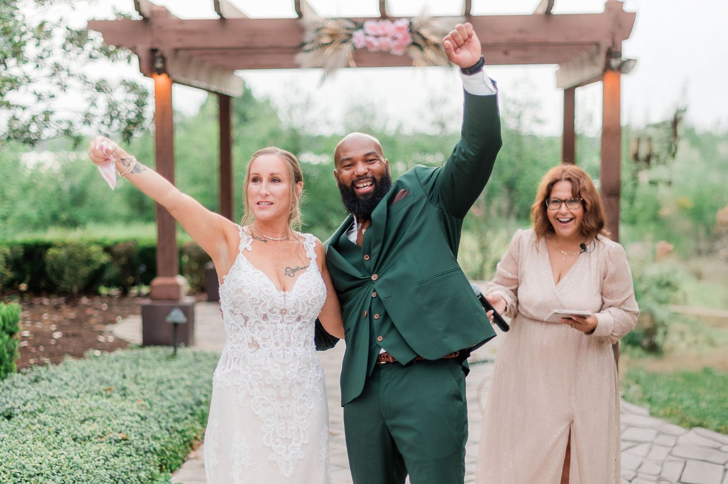 Wedding couple and officiant celebrate with raised arms under wooden pergola.