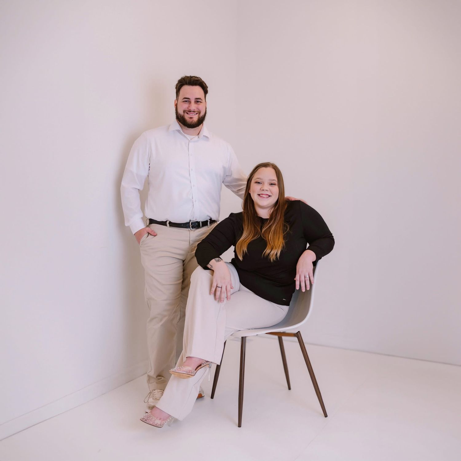 A man and woman pose professionally in a bright white studio, man standing and woman seated in a modern chair.