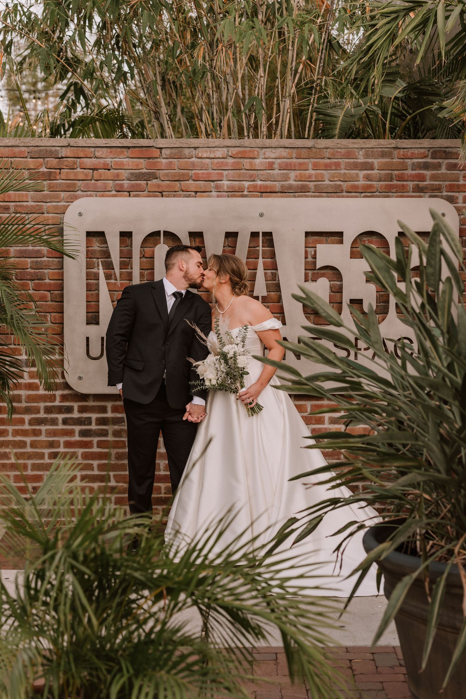 Elegant wedding couple posing against brick wall with tropical palm plants.