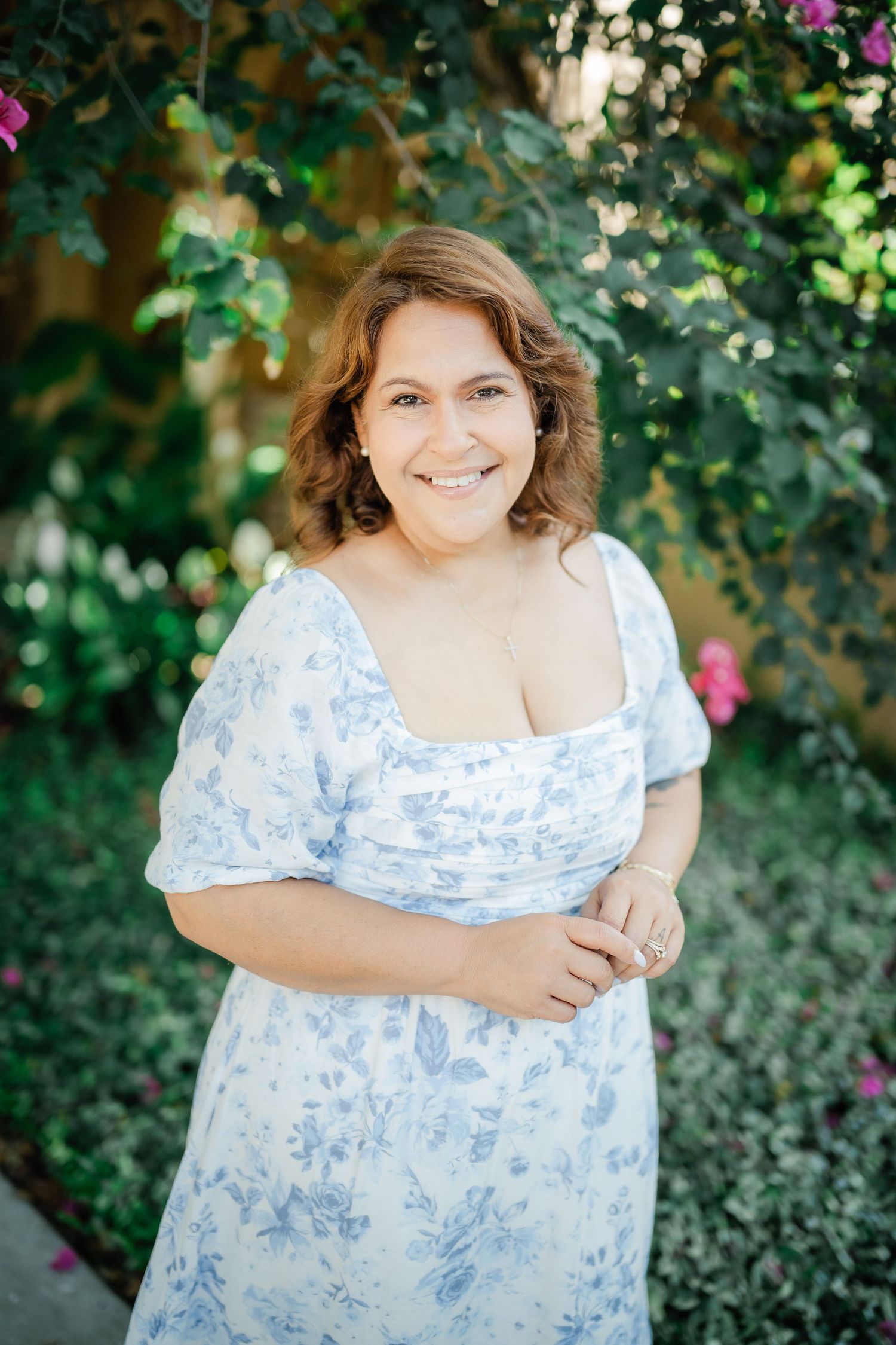 A person in a blue and white floral dress poses in a garden setting with blooming flowers.
