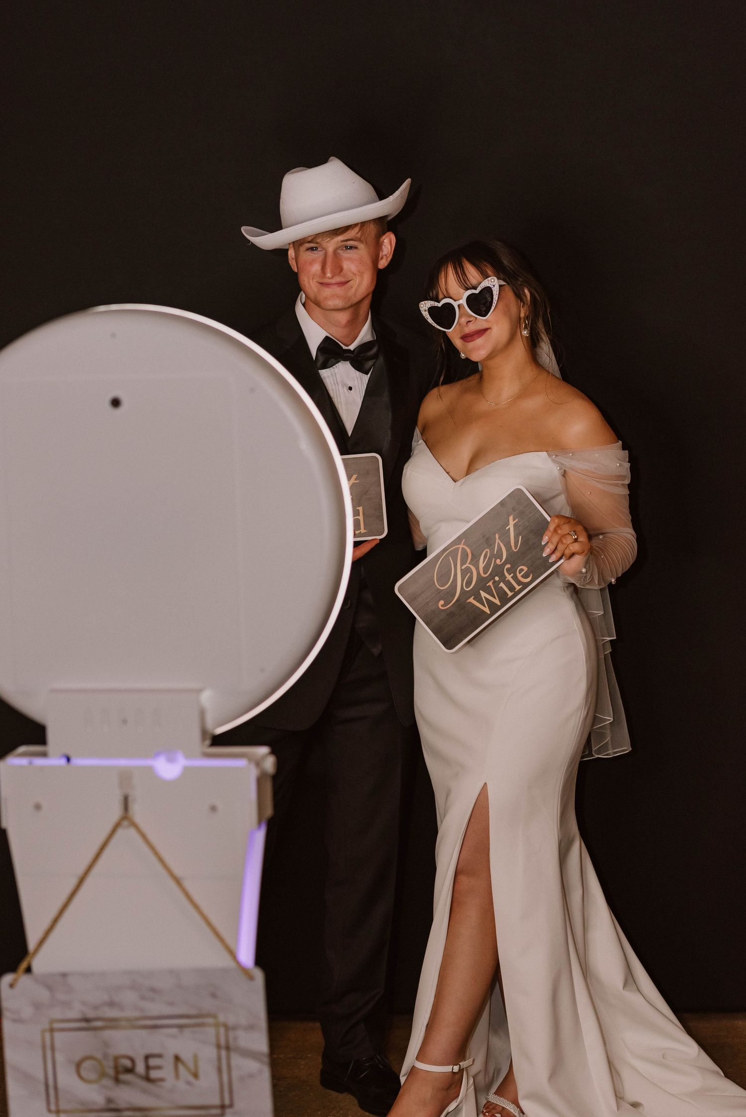 Photo booth scene with couple in formal attire and fun props against dark background.