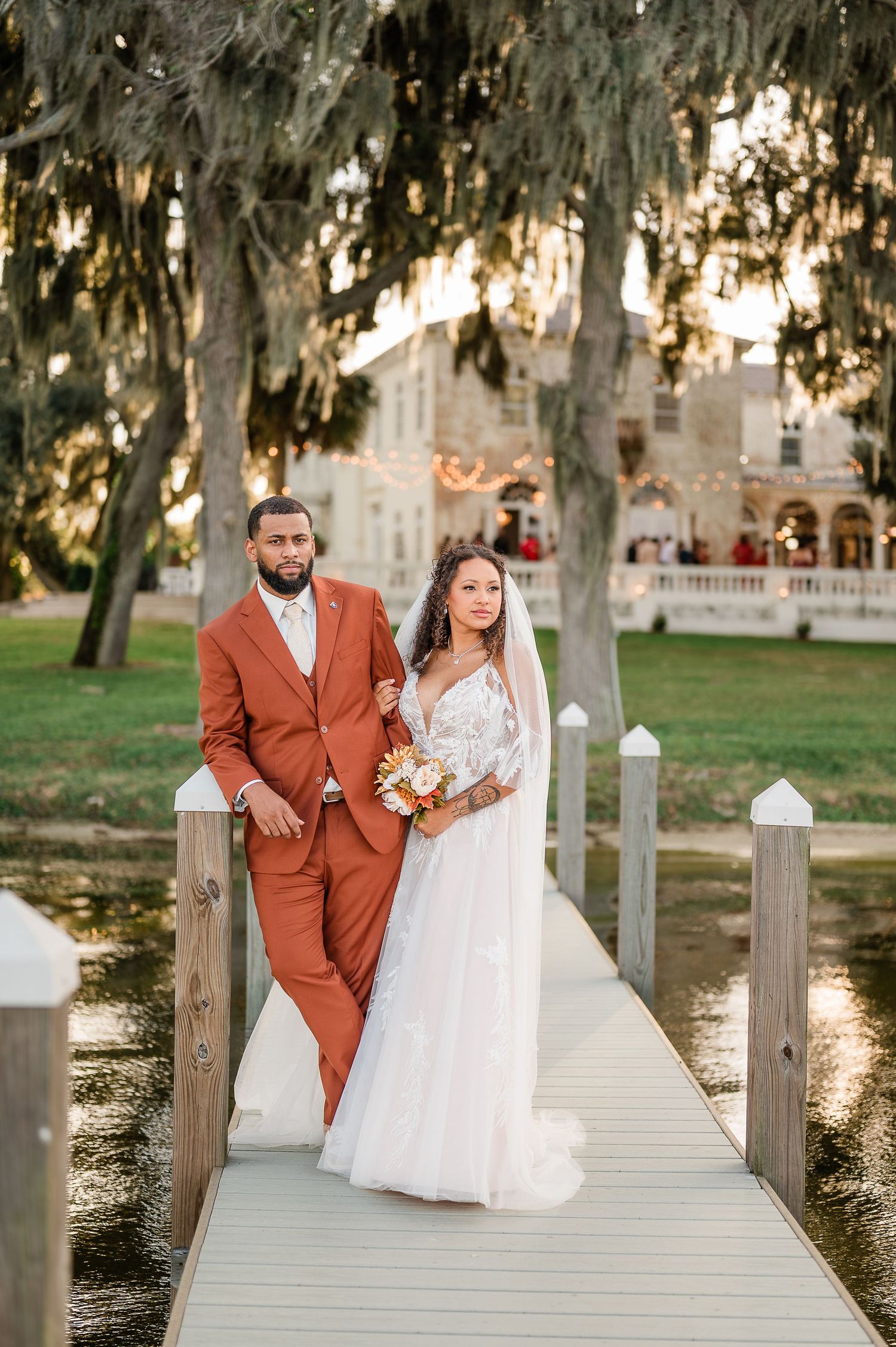 Newlywed couple poses on wooden dock at sunset wearing white dress and rust-colored suit.