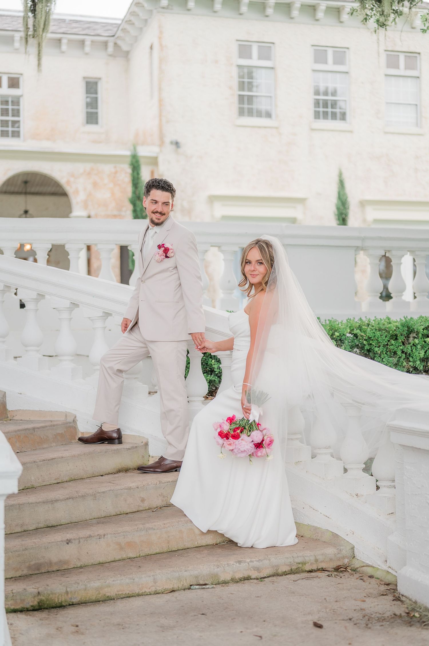 A wedding couple in white attire poses on elegant stone steps at a historic venue with white columns and arches.