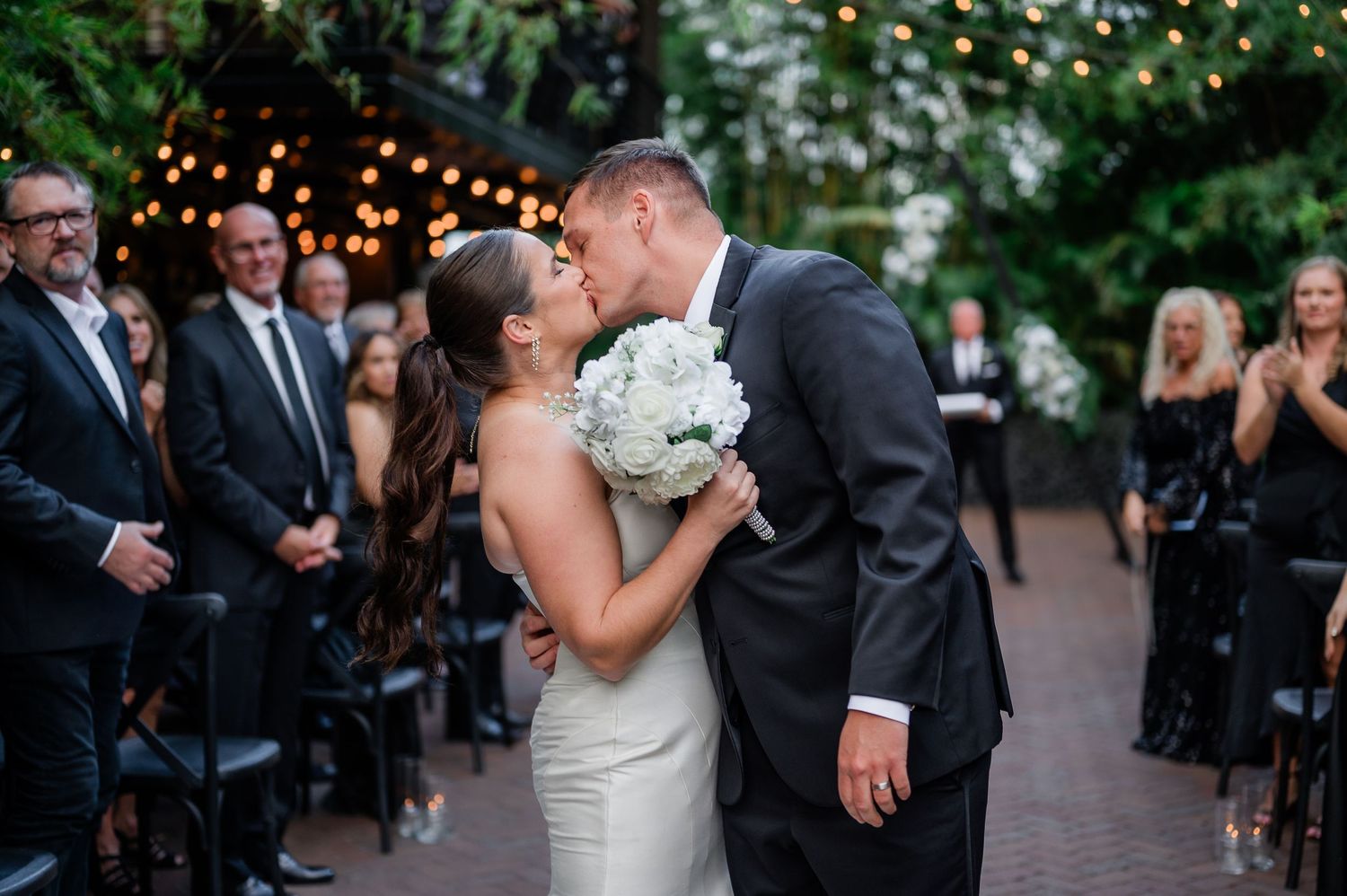 A couple shares a romantic kiss during an outdoor wedding ceremony with guests watching in the background.