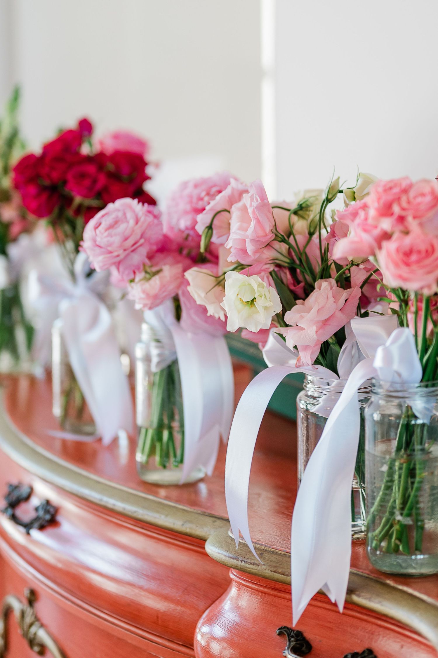 Small glass jars with pink roses and white ribbons line up elegantly on a rustic wooden surface.