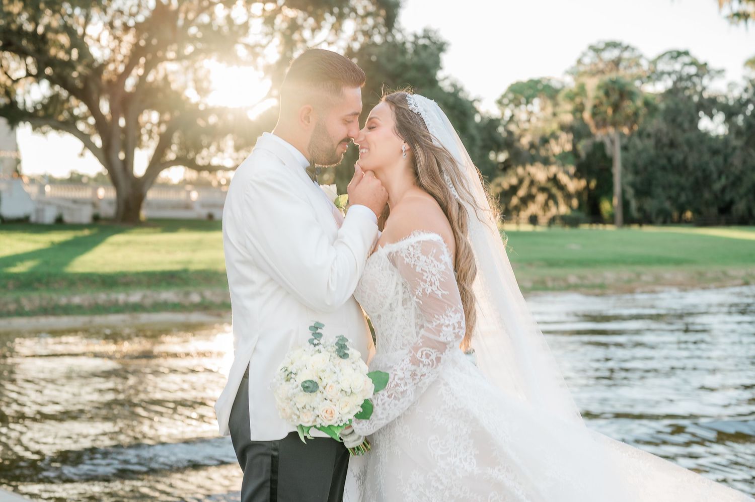 A couple in wedding attire embraces by a scenic lake during golden hour sunset.