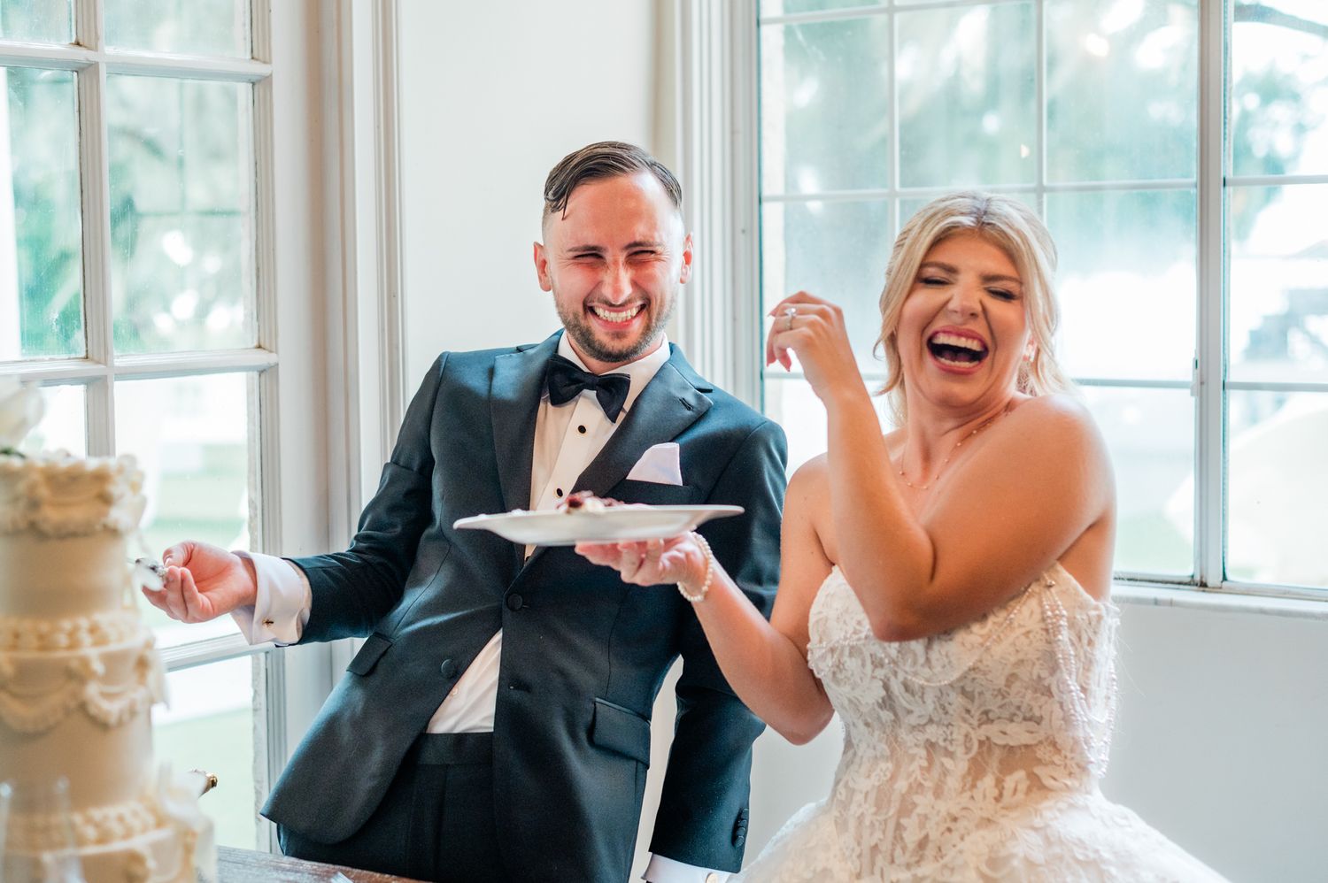 Bride and groom laughing while cutting wedding cake, groom in navy tuxedo, bride in beaded gown.