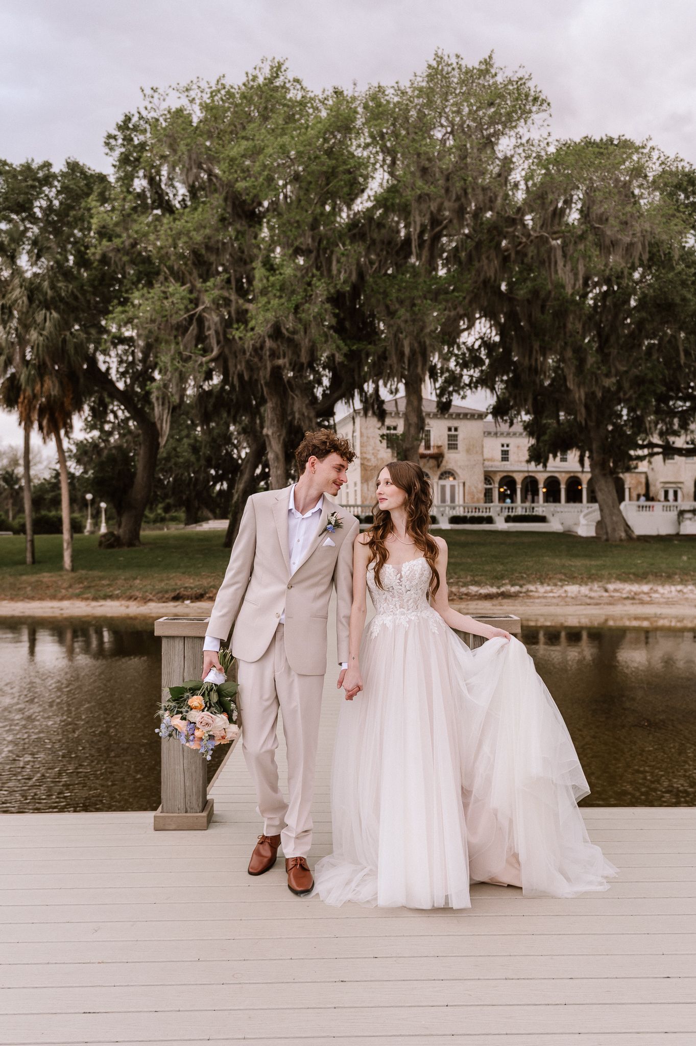 Couple in wedding attire pose on dock with Spanish moss trees and mansion in background.