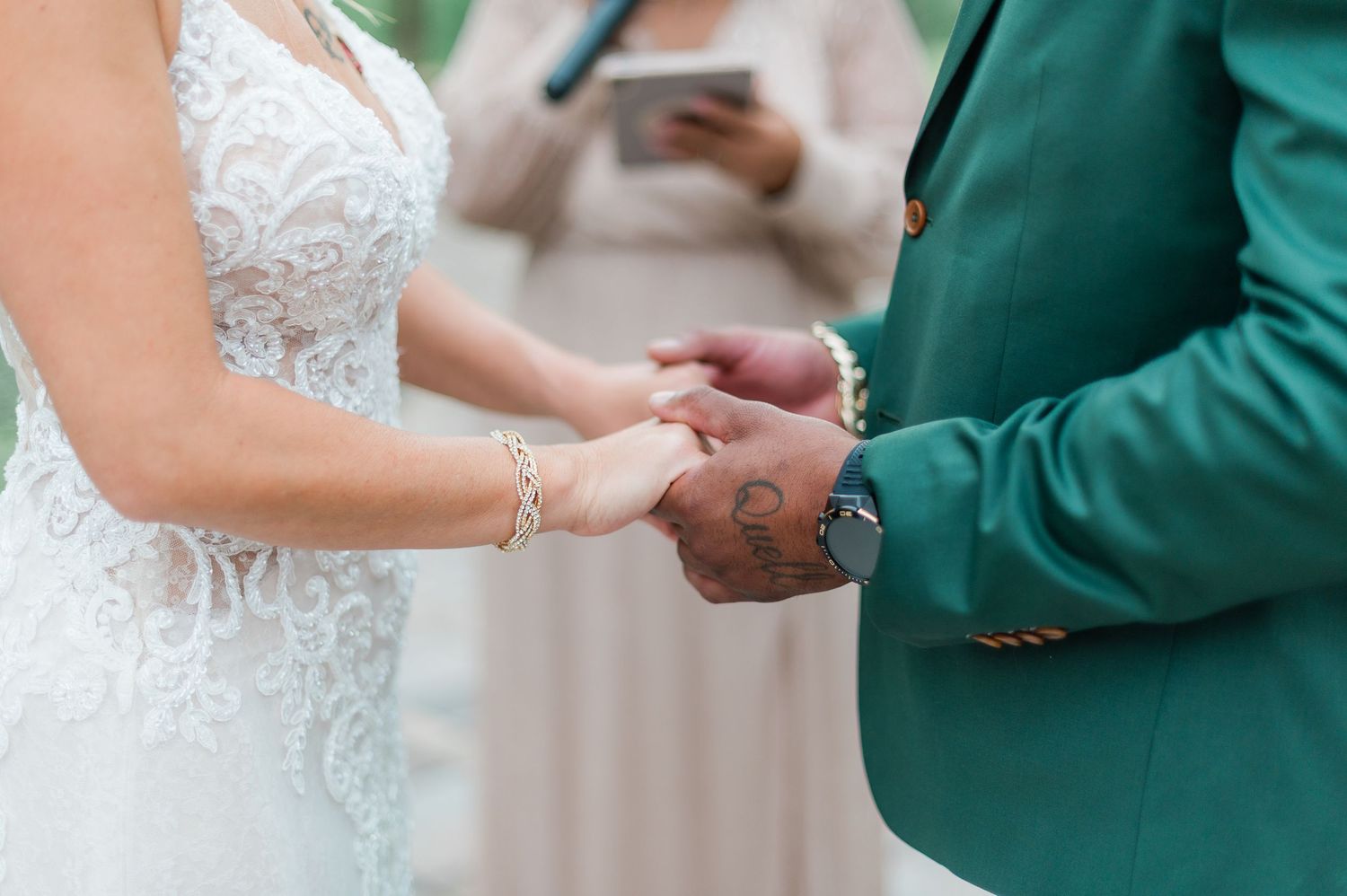 Close-up of wedding couple holding hands during ceremony showing lace dress and green suit details.