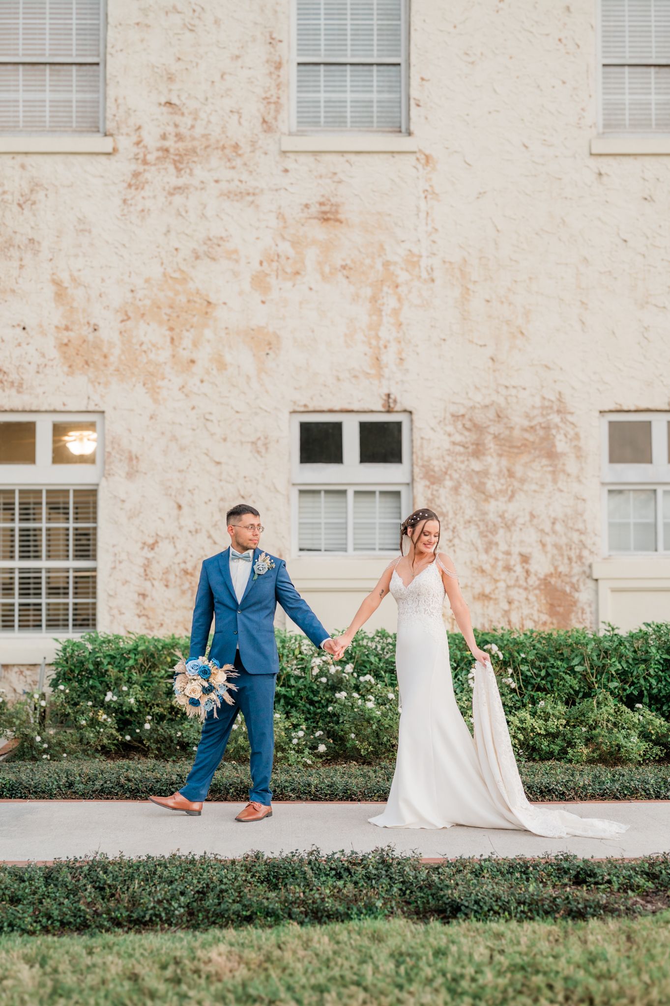 A couple in wedding attire playfully poses on a grassy lawn in front of a historic building exterior.