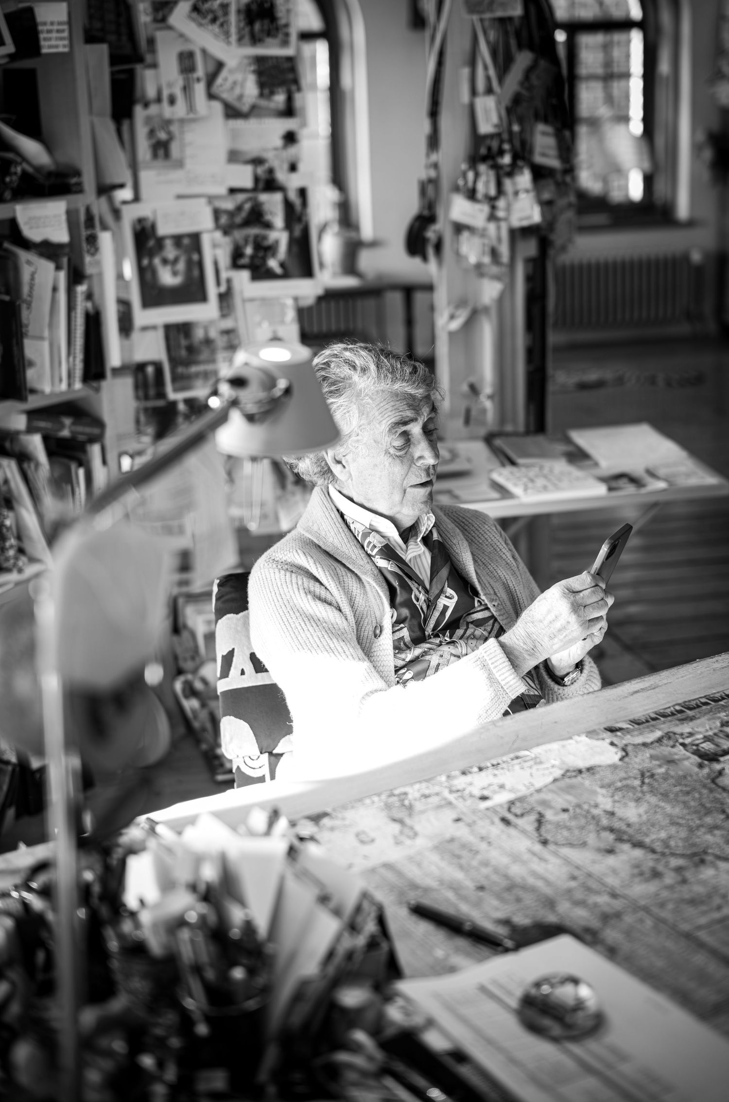 Black and white photo of someone working at a desk in a studio with photos and artwork displayed on the wall.
