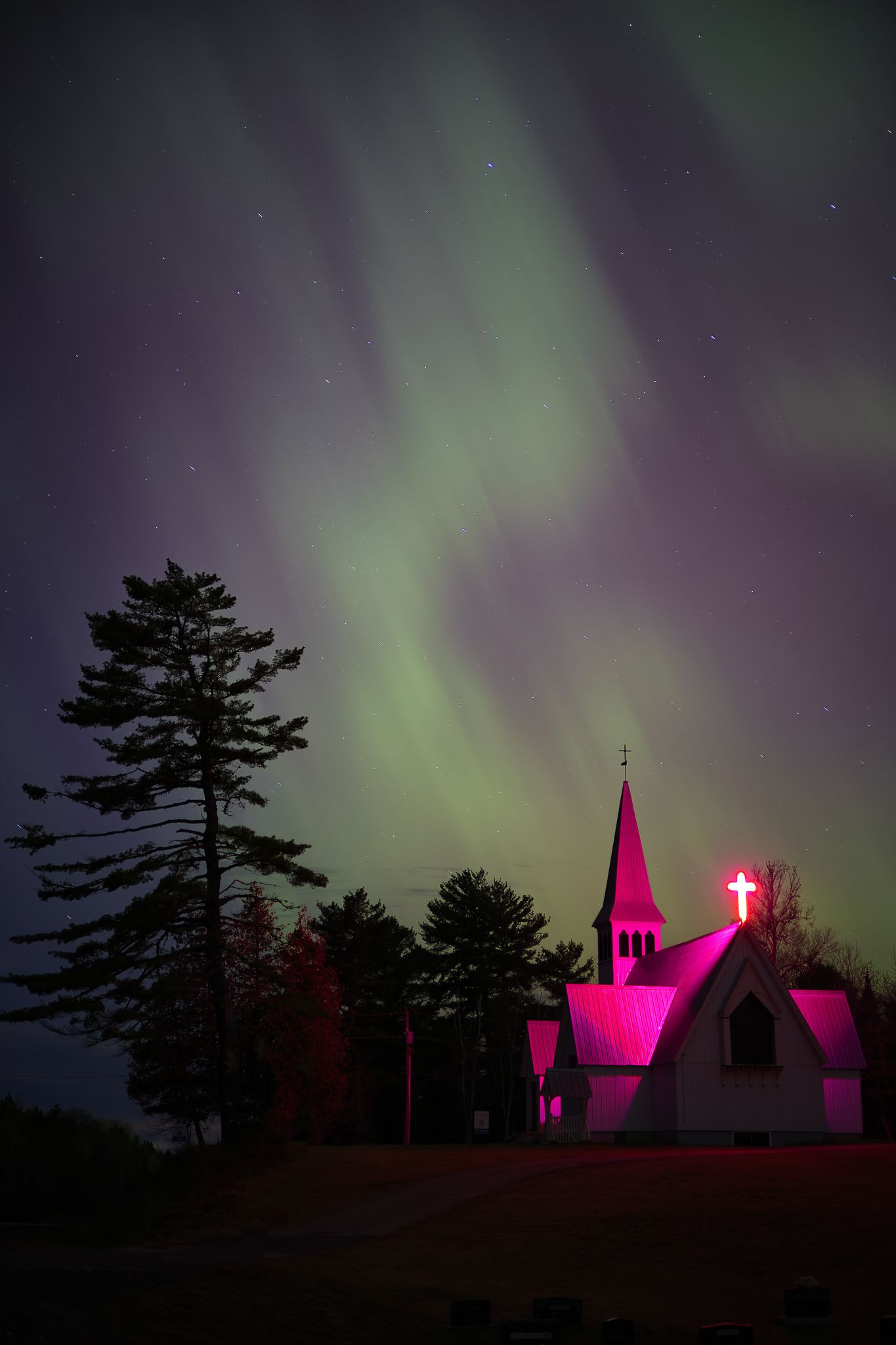 Silhouette of pink-lit church steeples against a backdrop of green aurora borealis dancing in the night sky, In New Brunswick