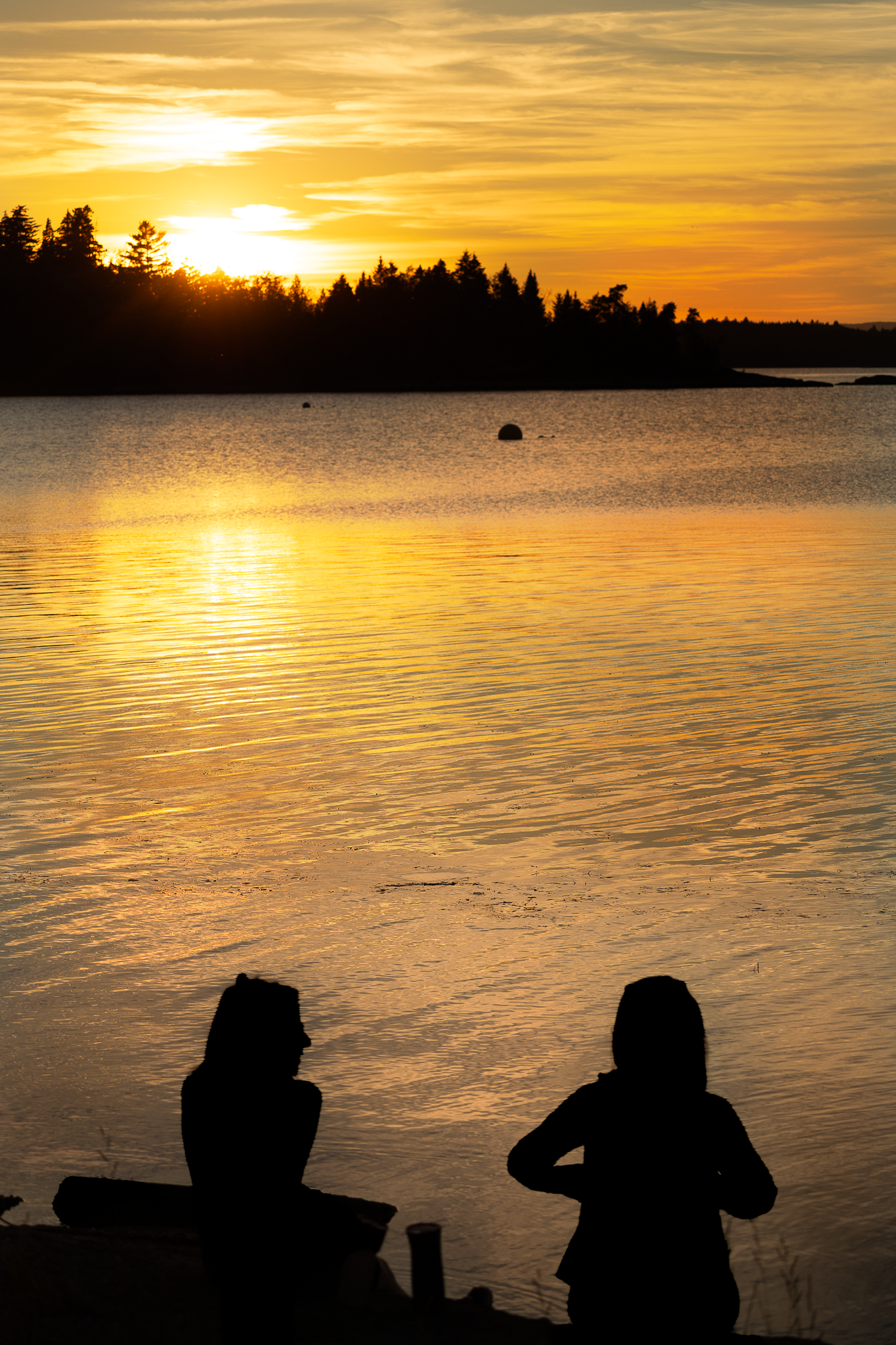 Silhouettes of people sit along a lake shore at sunset as golden light reflects off the calm water in Saint John.