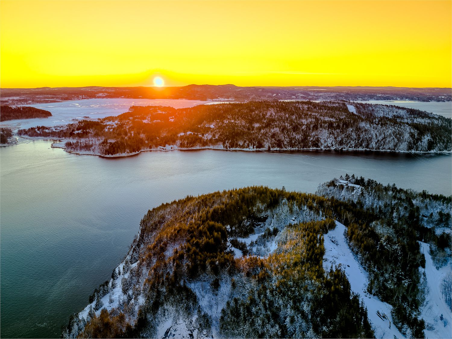 Aerial panoramic view of a snow-covered coastline at sunset with golden light reflecting off the water and rocky cliffs., Saint John River.