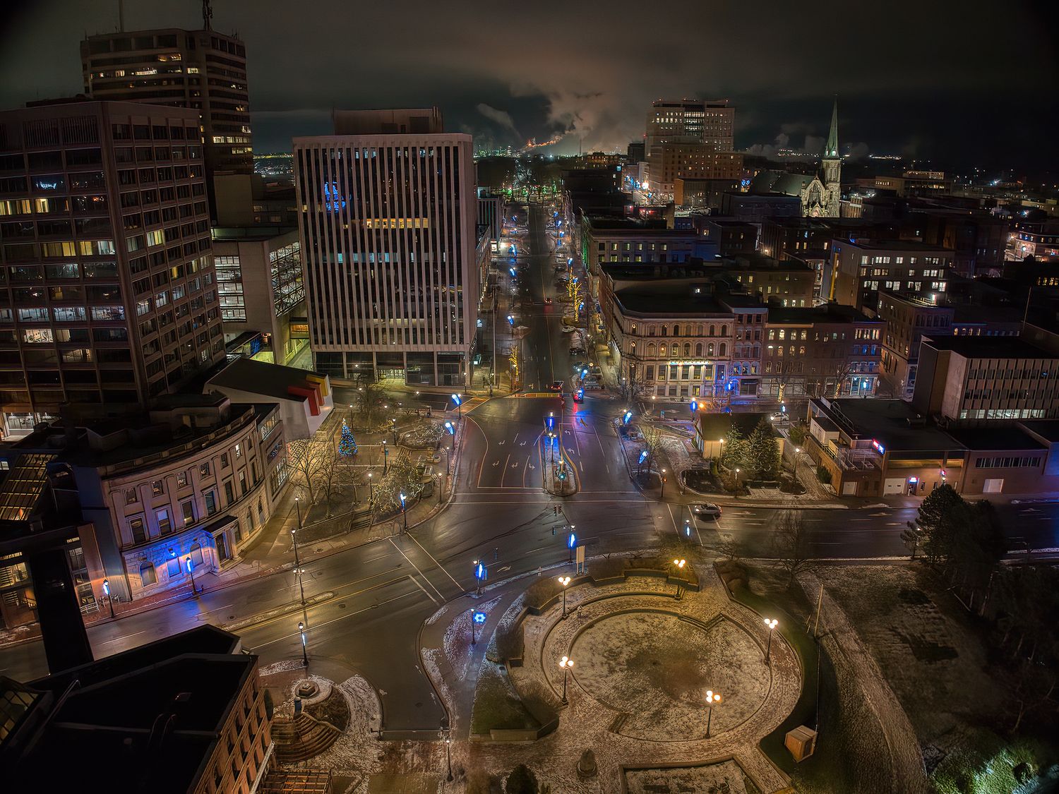 Aerial nighttime view of illuminated city streets and buildings with colorful lights reflecting on wet pavement, lower King St. Saint John.