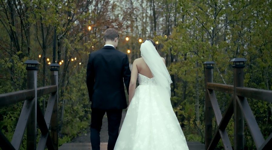 A couple in wedding attire walks across a wooden bridge decorated with string lights at dusk in Moncton.