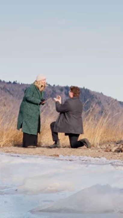 Two people in winter coats photographing something on a snowy shoreline with mountains in the background.