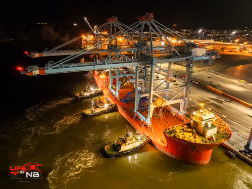 An aerial drone shot at night, featuring 3 tugboats pushing a cargo ship with cranes into the dock, being delivered to Saint John, NB.