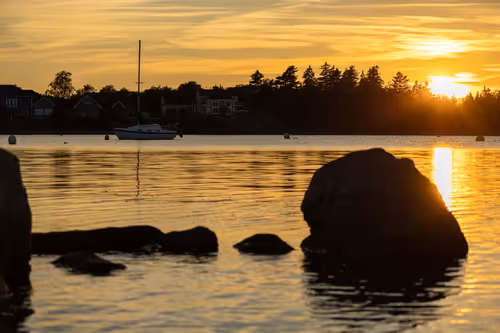 A golden hour shot featuring a sailboat in background, golden light reflecting off the water, silhouette rocks in foreground, Saint John NB