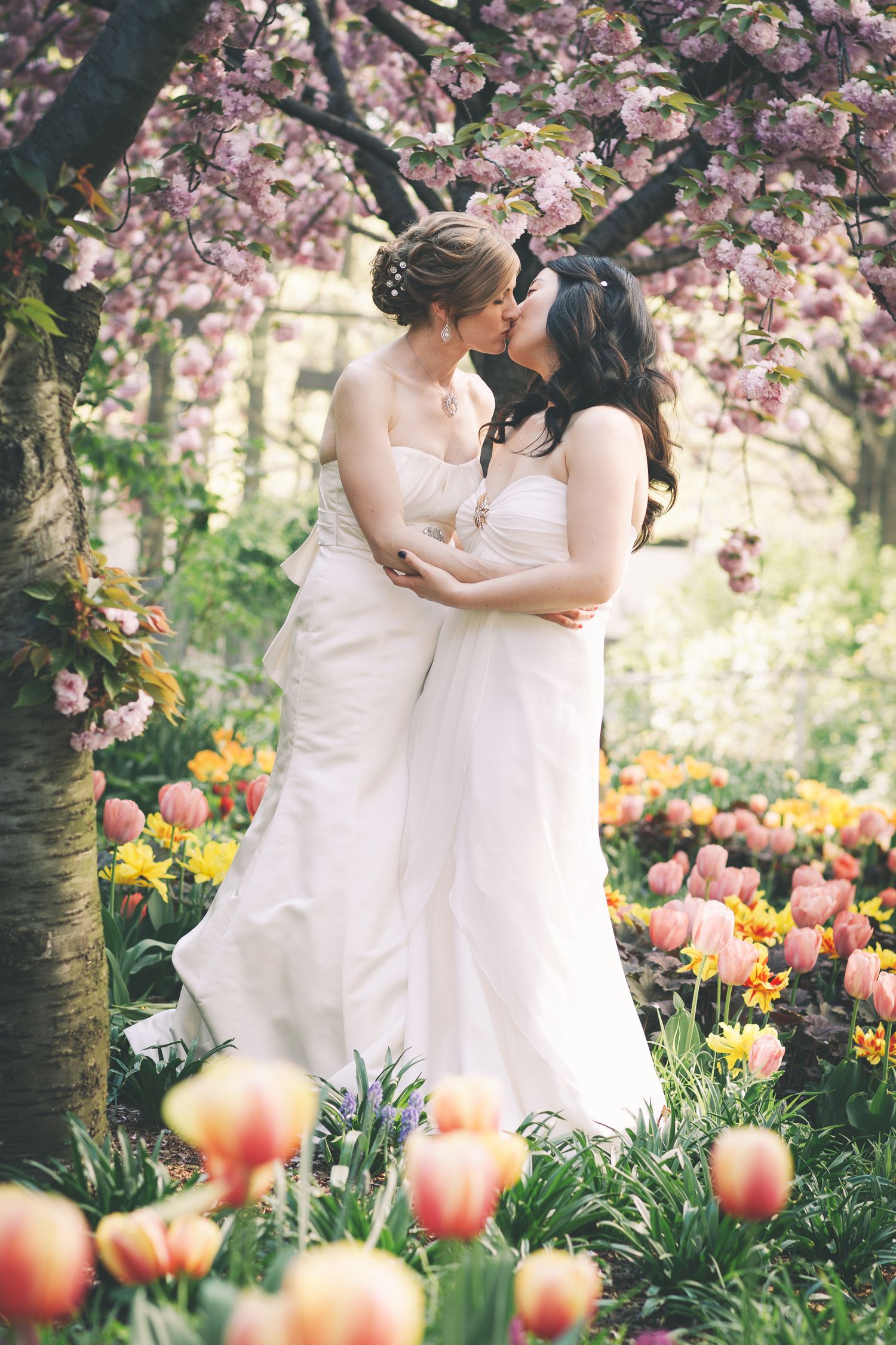Two brides in white dresses embrace under cherry blossoms surrounded by spring tulips. candid wedding photography Gold Coast