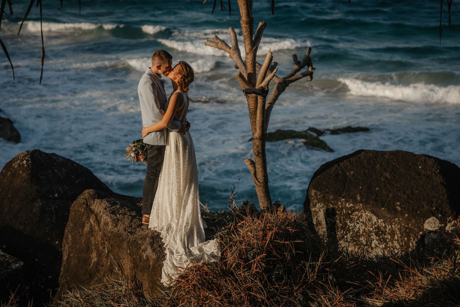 A couple embraces on rocky coastal cliffs with ocean waves crashing behind them. Gold Coast beach wedding photography. Froggies beach