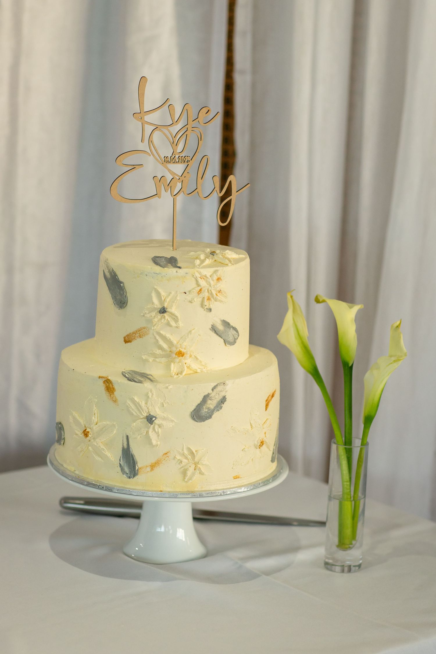 Two-tier white wedding cake with floral details and gold name topper beside calla lilies. beach wedding currumbin surf club wedding