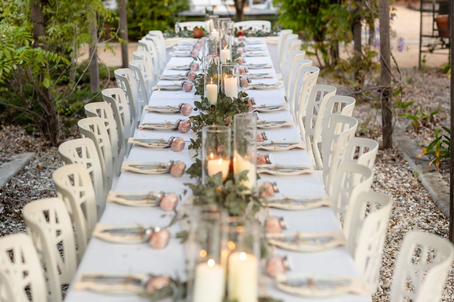 Long elegant table setting with white linens and place settings under trees for an outdoor reception.