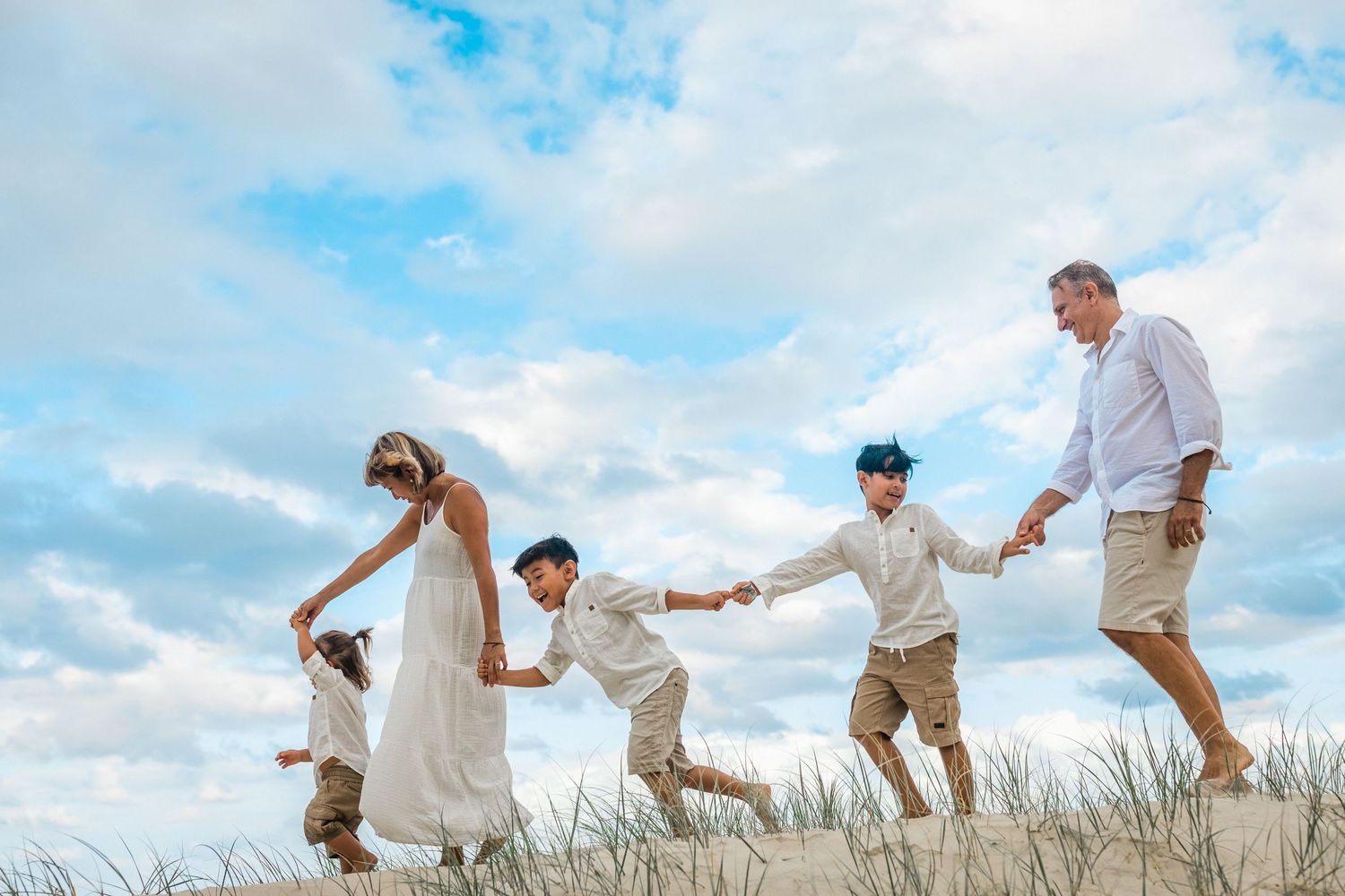 Family plays together while running through sand dunes against blue sky at sunset.Gold Coast candid family photographer.