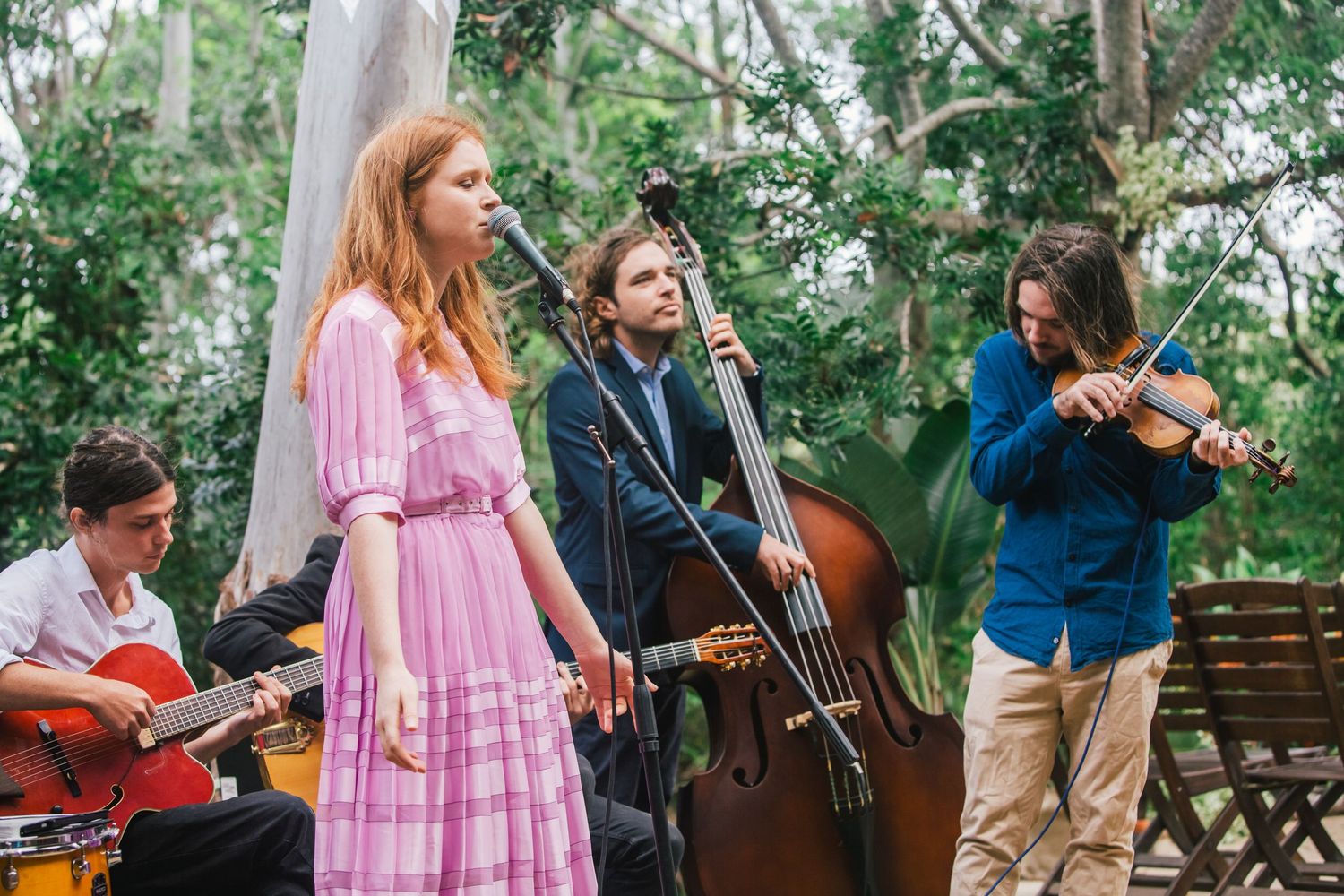 Musicians playing instruments outdoors with lush greenery in the background.