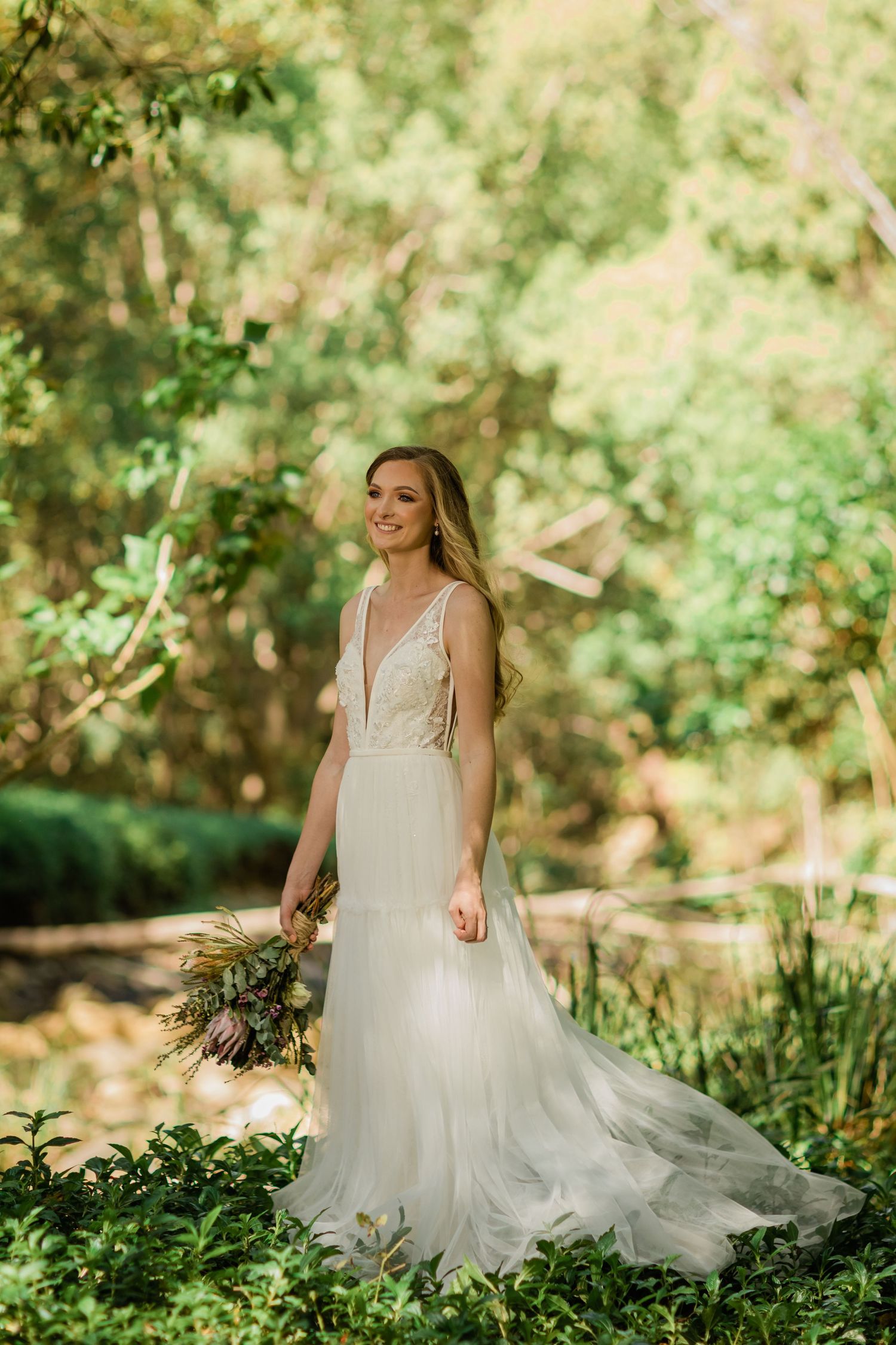 A bride in a flowing white gown poses gracefully in a sunlit garden setting with lush greenery.Gold Coast beach wedding photography