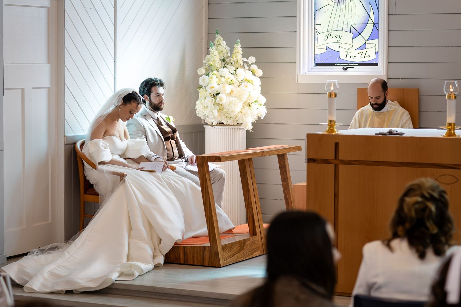 A bride in a white gown sits on a wooden church pew near stained glass windows.Gold Coast hinterland wedding photographer