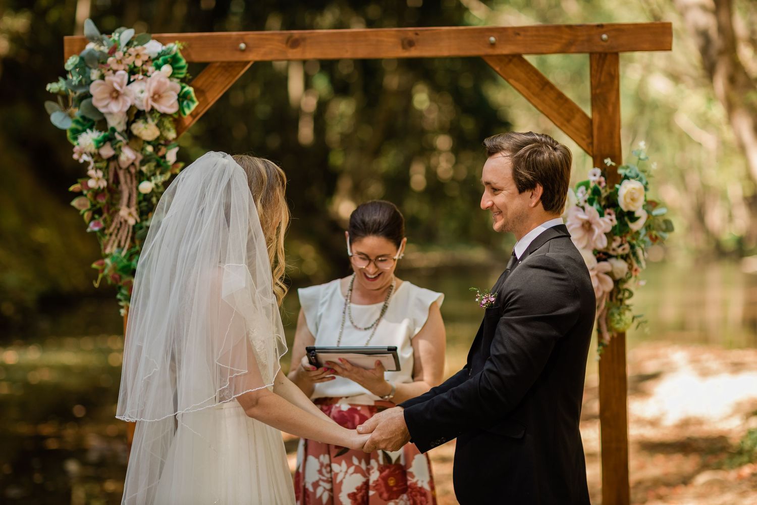 Wedding ceremony takes place under a wooden arch decorated with flowers in a forest setting.Currumbin Valley wedding photography Gold Coast