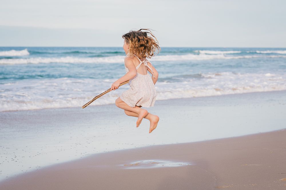 A carefree child jumps joyfully on a sandy beach while holding a stick, with ocean waves in the background.