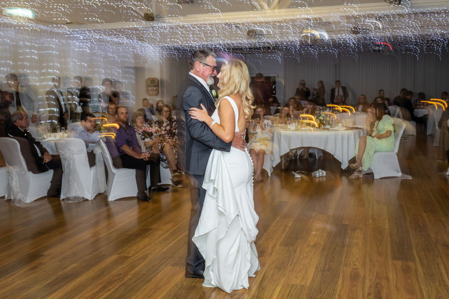 A couple shares their first dance at a wedding reception on a polished wooden dance floor surrounded by decorated tables.